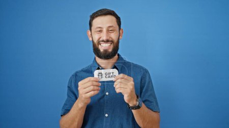 Foto de Joven hombre hispano sonriendo confiado sosteniendo tarjeta de regalo sobre fondo azul aislado - Imagen libre de derechos