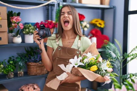 Photo for Young brunette woman working at florist shop holding camera angry and mad screaming frustrated and furious, shouting with anger looking up. - Royalty Free Image