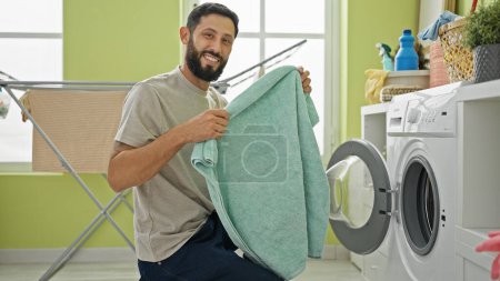 Téléchargez les photos : Jeune homme hispanique laver des vêtements tenant serviette souriant à la buanderie - en image libre de droit