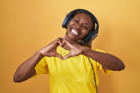 Téléchargez les photos : Femme afro-américaine écoutant de la musique à l'aide d'écouteurs souriant dans l'amour faire forme de symbole de coeur avec les mains. concept romantique. - en image libre de droit
