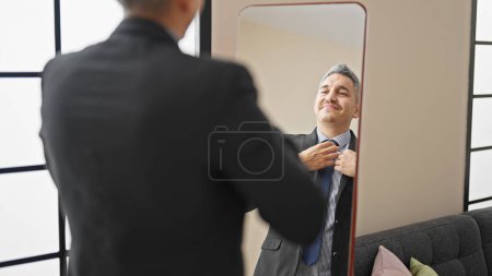 Photo for Young hispanic man business worker wearing suit looking on mirror at home - Royalty Free Image