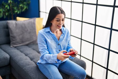 Photo for Young beautiful hispanic woman playing video game sitting on sofa at home - Royalty Free Image