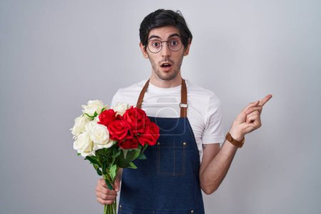 Foto de Joven hombre hispano sosteniendo ramo de rosas blancas y rojas sorprendido señalando con el dedo hacia un lado, boca abierta expresión asombrada. - Imagen libre de derechos
