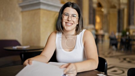Téléchargez les photos : Jeune belle femme hispanique portant des lunettes souriant heureux menu de lecture à la cafétéria - en image libre de droit