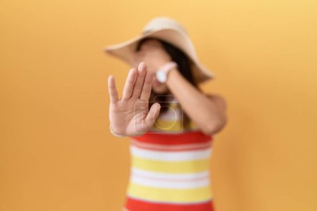 Foto de Mujer china de mediana edad con sombrero de verano sobre fondo amarillo cubriendo los ojos con las manos y haciendo gesto de parada con expresión triste y miedo. concepto avergonzado y negativo. - Imagen libre de derechos