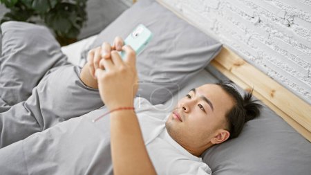 Photo for Handsome young chinese man, in relaxed fashion, lying comfortably in bed at home using smartphone in bedroom, immersed in a serious online world - Royalty Free Image