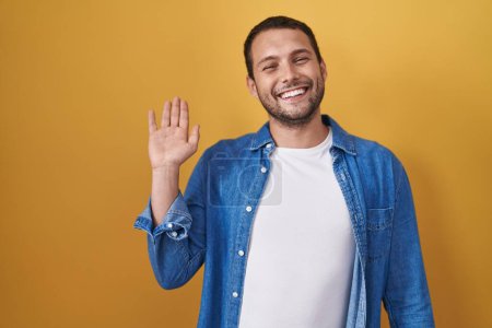 Téléchargez les photos : Homme hispanique debout sur fond jaune renonçant à dire bonjour heureux et souriant, geste de bienvenue amical - en image libre de droit
