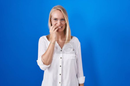 Foto de Young caucasian woman standing over blue background looking stressed and nervous with hands on mouth biting nails. anxiety problem. - Imagen libre de derechos