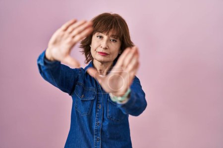 Téléchargez les photos : Femme d'âge moyen debout sur fond rose faisant cadre en utilisant les mains paumes et les doigts, perspective de la caméra - en image libre de droit