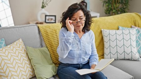 Photo for Middle age hispanic woman reading book sitting on sofa at home - Royalty Free Image