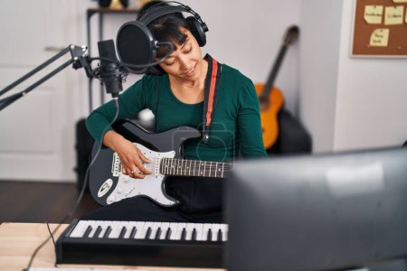 Téléchargez les photos : Jeune belle musicienne hispanique souriante confiante jouant de la guitare électrique au studio de musique - en image libre de droit