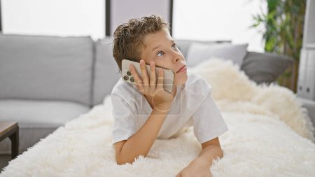 Photo for Adorable blond boy, deeply engrossed in a serious phone conversation, lies relaxed on a sofa at home. absolutely cute expression on his face using the latest technology indoors. - Royalty Free Image