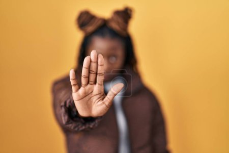 Foto de Mujer africana con el pelo trenzado de pie sobre fondo amarillo haciendo dejar de cantar con la palma de la mano. expresión de advertencia con gesto negativo y grave en la cara. - Imagen libre de derechos