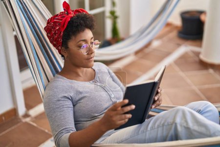 Photo for African american woman reading book lying on hammock at home terrace - Royalty Free Image