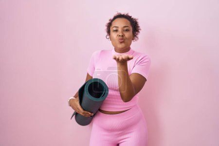 Foto de Mujer hispana joven con el pelo rizado sosteniendo estera de yoga sobre fondo rosa mirando a la cámara soplando un beso con la mano en el aire siendo encantadora y sexy. expresión de amor. - Imagen libre de derechos