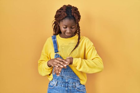 Téléchargez les photos : Femme africaine debout sur fond jaune vérifier l'heure sur montre-bracelet, détendu et confiant - en image libre de droit