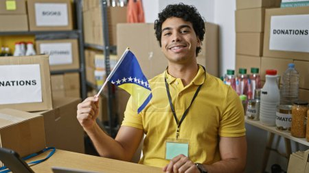 Téléchargez les photos : Jeune homme latin volontaire tenant bosnia et drapeau de l'herzénie souriant au centre de charité - en image libre de droit