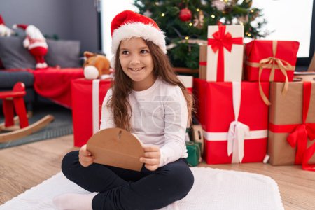 Téléchargez les photos : Adorable hispanique fille tenant santa claus lettre assis sur le sol par arbre de Noël à la maison - en image libre de droit