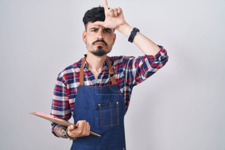 Téléchargez les photos : Jeune homme hispanique avec barbe portant tablier de serveur tenant presse-papiers se moquant des gens avec les doigts sur le front faisant geste de perdant moqueur et insultant. - en image libre de droit