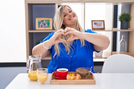 Téléchargez les photos : Femme blanche plus la taille de manger le petit déjeuner à la maison souriant dans l'amour faisant forme de symbole de coeur avec les mains. concept romantique. - en image libre de droit