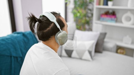 Photo for Handsome young chinese man, embracing a relaxed lifestyle, sitting back on his home sofa, blissfully lost in the captivating song, listening through his cherished gadget - headphones - Royalty Free Image
