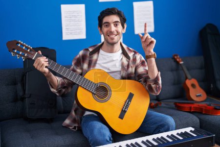 Téléchargez les photos : Young hispanic man playing classic guitar at music studio surprised with an idea or question pointing finger with happy face, number one - en image libre de droit