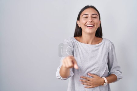 Foto de Mujer hispana joven de pie sobre fondo blanco riéndose de ti, señalando con el dedo a la cámara con la mano sobre el cuerpo, expresión de vergüenza - Imagen libre de derechos