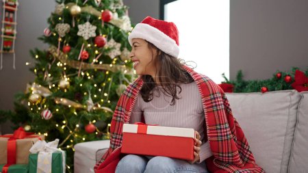 Téléchargez les photos : Young beautiful hispanic woman unpacking gift sitting by christmas tree at home - en image libre de droit