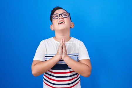 Téléchargez les photos : Jeune enfant hispanique debout sur fond bleu mendier et prier avec les mains ainsi que l'expression d'espoir sur le visage très émotionnel et inquiet. mendier. - en image libre de droit