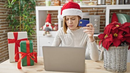 Téléchargez les photos : Jeune femme blonde faisant du shopping avec ordinateur portable et carte de crédit célébrant Noël regardant bouleversé à la salle à manger - en image libre de droit