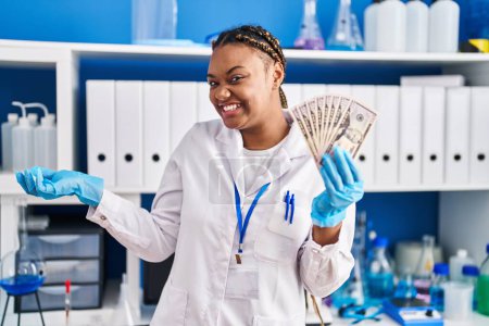 Photo for African american woman with braids working at scientist laboratory holding money screaming proud, celebrating victory and success very excited with raised arm - Royalty Free Image