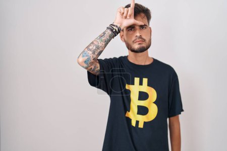 Téléchargez les photos : Jeune homme hispanique avec des tatouages portant Bitcoin t-shirt se moquant des gens avec les doigts sur le front faisant geste perdant moquerie et insultant. - en image libre de droit