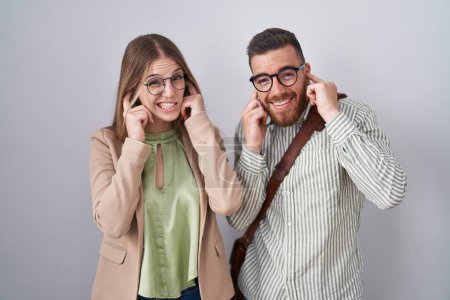 Téléchargez les photos : Jeune couple debout sur fond blanc couvrant les oreilles avec des doigts avec une expression agacée pour le bruit de la musique forte. concept sourd. - en image libre de droit