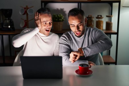 Téléchargez les photos : Couple homosexuel à l'aide d'un ordinateur portable en regardant le temps de la montre inquiet, peur d'arriver en retard - en image libre de droit