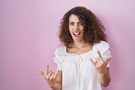 Photo for Hispanic woman with curly hair standing over pink background shouting with crazy expression doing rock symbol with hands up. music star. heavy concept. - Royalty Free Image