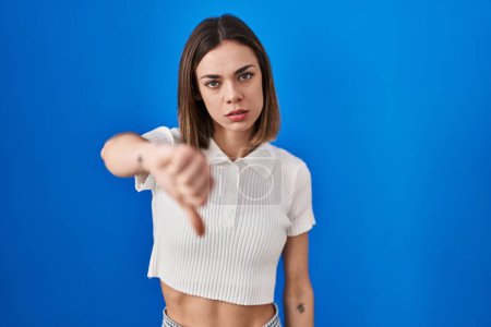 Photo for Hispanic woman standing over blue background looking unhappy and angry showing rejection and negative with thumbs down gesture. bad expression. - Royalty Free Image