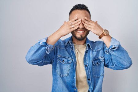 Foto de Joven hombre hispano de pie sobre un fondo aislado cubriendo los ojos con las manos sonriendo alegre y divertido. concepto ciego. - Imagen libre de derechos