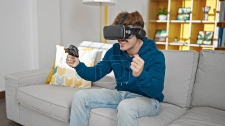 Téléchargez les photos : Jeune homme hispanique jouer à un jeu vidéo en utilisant joystick et lunettes de réalité virtuelle à la maison - en image libre de droit