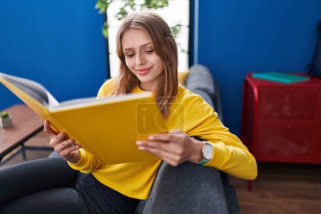 Téléchargez les photos : Young blonde woman reading book sitting on sofa at home - en image libre de droit