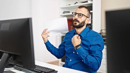 Téléchargez les photos : Jeune homme hispanique travailleur d'affaires utilisant l'ordinateur en utilisant des documents comme ventilateur de main au bureau - en image libre de droit