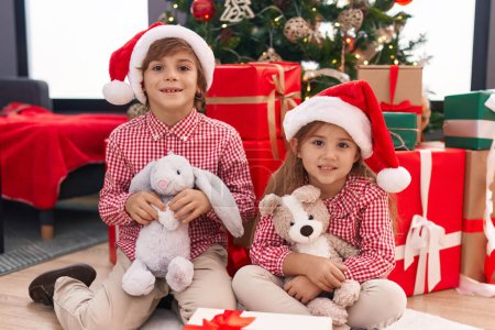 Photo for Brother and sister holding teddy bear sitting on floor by christmas tree at home - Royalty Free Image