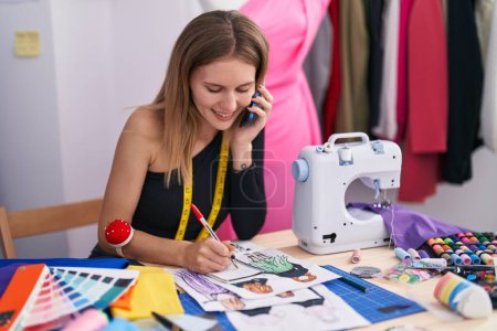Téléchargez les photos : Jeune femme blonde tailleur parler sur smartphone dessin conception de vêtements au magasin de tailleur - en image libre de droit