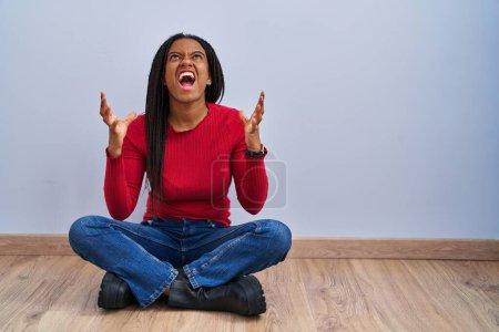 Foto de Joven afroamericano con trenzas sentado en el suelo en casa loco y loco gritando y gritando con expresión agresiva y los brazos levantados. concepto de frustración. - Imagen libre de derechos