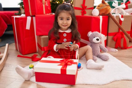 Téléchargez les photos : Adorable fille hispanique assise sur le sol par des cadeaux de Noël jouant à la maison - en image libre de droit