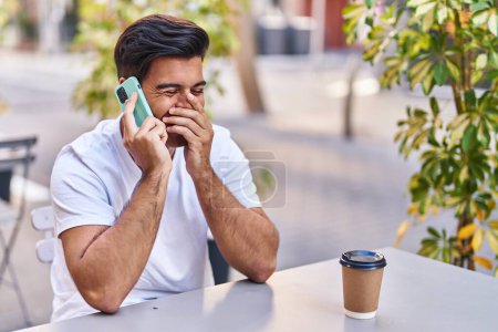 Téléchargez les photos : Jeune homme hispanique parlant sur smartphone boire du café à la terrasse du café - en image libre de droit