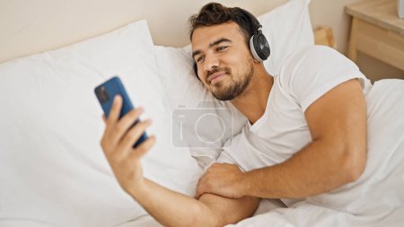 Foto de Joven hombre hispano viendo video en smartphone acostado en la cama en el dormitorio - Imagen libre de derechos