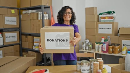 Téléchargez les photos : Moyen Age hispanique femme bénévole souriant confiant tenant paquet de dons au centre de charité - en image libre de droit