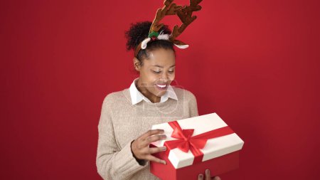 Foto de Mujer afroamericana desempacando regalo de Navidad con orejas de reno sobre fondo rojo aislado - Imagen libre de derechos