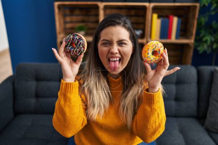 Téléchargez les photos : Jeune femme hispanique mangeant des beignets à la maison langue collante heureux avec une expression drôle. - en image libre de droit