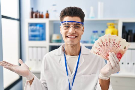 Photo for Young hispanic man working at scientist laboratory holding shekels celebrating achievement with happy smile and winner expression with raised hand - Royalty Free Image